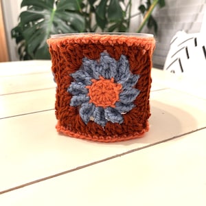 May include: A handmade crochet cup cozy in shades of brown, orange, and blue. The cozy features a square design with a large flower in the center. The flower has blue petals and an orange center. The cozy is designed to fit around a standard-sized cup.