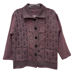 Puede incluir: Una chaqueta de punto de color morado con un dise&ntilde;o floral negro y rosa. La chaqueta tiene un cierre de botones y dos bolsillos.