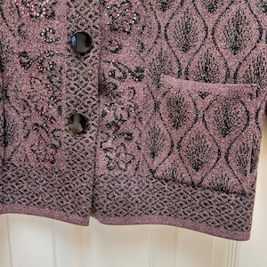 Puede incluir: Primer plano de una chaqueta de punto morada con patrones florales negros y rosas y botones negros. La chaqueta tiene un bolsillo en el lado derecho.