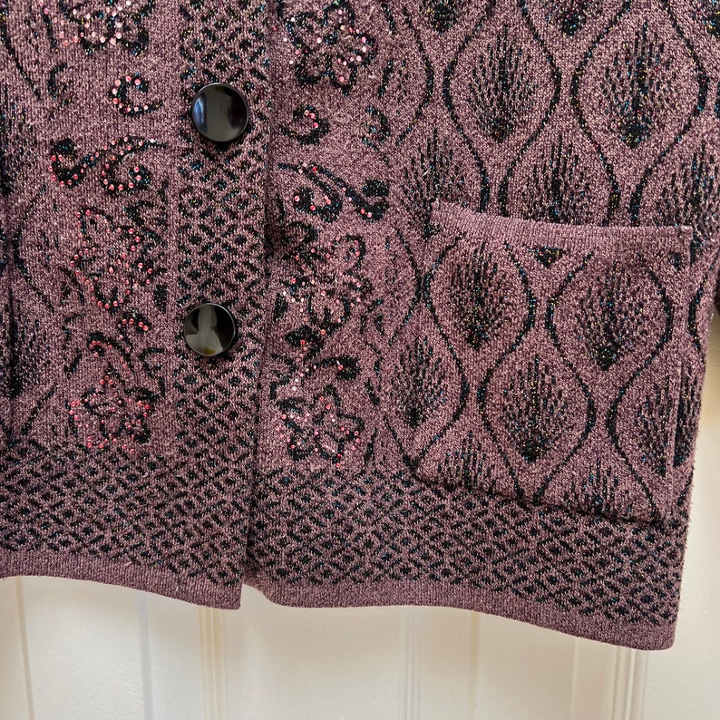 Puede incluir: Primer plano de una chaqueta de punto morada con patrones florales negros y rosas y botones negros. La chaqueta tiene un bolsillo en el lado derecho.