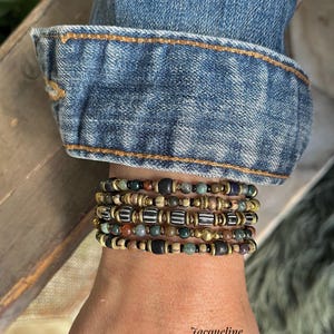 Puede incluir: Un conjunto de pulseras de cuentas en varios colores, incluyendo negro, marrón, verde y dorado, se lleva en una muñeca. Las pulseras están hechas de diferentes tipos de cuentas. Se ve una manga de chaqueta vaquera. El texto "Jacqueline Koster Sieraden" está en la parte inferior.