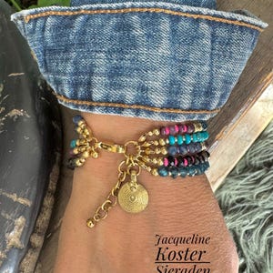 Op de afbeelding: Een meerrijige kralenarmband met rijen kleine, gefacetteerde kralen in magenta, turquoise, blauw en zwart, afgewisseld met goudkleurige metalen kralen. Een goudkleurige ronde bedel met een spiraalpatroon hangt aan de sluiting.
