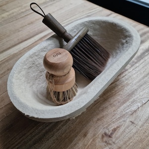 Op de afbeelding: Een witte keramische kom met twee houten borstels. Eén borstel heeft een houten handvat met het woord "bio" erop gedrukt. De andere borstel heeft een houten handvat met het woord "olo" erop gedrukt.