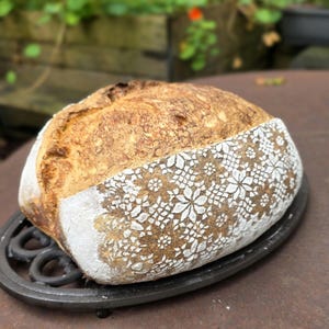 Pode incluir: Um pão de massa madre recém-assado com um padrão floral branco decorativo em um lado. A crosta é marrom dourado e o pão repousa sobre um trivet de ferro fundido preto. A imagem é tirada ao ar livre.