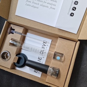 Pode incluir: Uma caixa de cartão aberta contendo um conjunto de arejador de vinho. O conjunto inclui um arejador preto e transparente, tubos de vidro com marcações de medição e um anel de vidro transparente. O texto "BILLIE OLIVE" é visível.