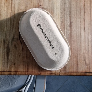 Puede incluir: Un recipiente de papel blanco de forma ovalada con el texto "@yournamehere b/o" impreso en él. El recipiente está sobre una superficie de madera.