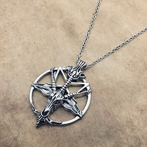 May include: Silver-coloured pendant necklace featuring a detailed Baphomet design within a pentagram. The pendant hangs from a delicate chain. The necklace is displayed on a neutral-toned background.
