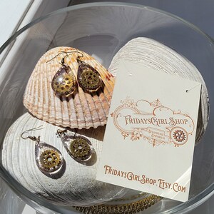 Peut inclure: Une paire de boucles d'oreilles en forme de goutte d'eau dorées et brunes avec un motif d'engrenage à l'intérieur. Les boucles d'oreilles sont présentées sur une coquille Saint-Jacques et une carte qui indique "FridaysGirlShop.Etsy.com".