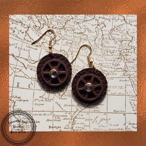 Peut inclure: Une paire de boucles d'oreilles en cuir marron avec un motif d'engrenage en cuivre et un strass transparent au centre de chaque engrenage. Les boucles d'oreilles sont suspendues à des crochets dorés.