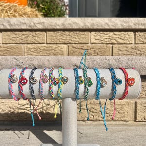 May include: A display of colorful braided bracelets with various charms, including evil eye and hamsa hand designs. The bracelets are displayed on a white stand.