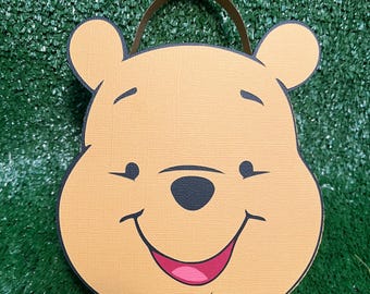 Puede incluir: Una bolsa de regalo redonda de color beige con el tema de Winnie the Pooh, con una cara sonriente, ojos negros y una boca rosa. La bolsa tiene un asa marrón y un contorno negro. El fondo es verde.