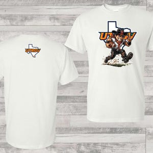 May include: White t-shirt with a cartoon cowboy running with a football, the text "UTRGV" and a Texas outline. The back of the shirt displays the UTRGV logo within a Texas outline. A casual tee.