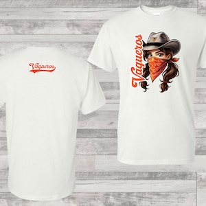 以下が含まれることがあります： バンダナとカウボーイハットをかぶったカウガールが描かれた白いTシャツ。前面にオレンジ色で縦に、背面に横に「Vaqueros」の文字がプリントされています。シャツは柔らかく快適な素材で作られています。