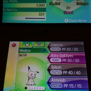 Pode incluir: Uma captura de tela de um jogo Nintendo 3DS mostrando um Pokémon Mimikyu de nível 10. O Pokémon é do tipo Fantasma e Fada com um fundo verde. O Pokémon tem um rosto rosa e branco com um nariz preto e olhos pretos. O Pokémon está segurando uma bola rosa e branca. Os movimentos do Pokémon são Copiar, Olhos de Boneca, Salpicar e Assustar. A habilidade do Pokémon é Disfarçar e ele está segurando uma Tag de Feitiço.