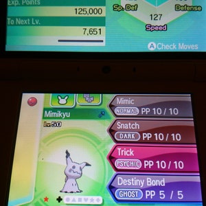 May include: A screenshot of a Nintendo DS game showing a Mimikyu Pokemon with the stats and moves of the Pokemon. The Pokemon is level 50 and has the moves Mimic, Snatch, Trick, and Destiny Bond.