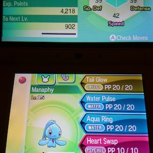 Può includere: Uno schermo mostra informazioni sul Pokémon Manaphy. La sezione superiore mostra statistiche come HP, Attacco e Velocità. La sezione inferiore mostra i movimenti come Tail Glow e Water Pulse. Il Pokémon è una piccola creatura blu.