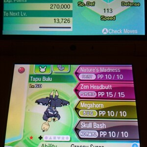 Può includere: Un dispositivo di gioco portatile mostra il profilo del Pokémon Tapu Bulu. Lo schermo mostra statistiche come HP, Attacco e Difesa, insieme alle mosse: Nature's Madness, Zen Headbutt, Megahorn e Skull Bash.