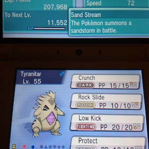 Pode incluir: Uma captura de tela de um videogame com um Tyranitar, um Pokémon do tipo Pedra e Sombrio. A tela mostra as estatísticas, movimentos e nível do Pokémon. A interface do jogo é visível, com texto em inglês e japonês.