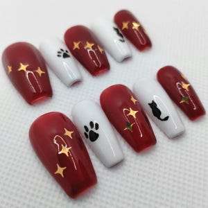 Op de afbeelding: Een set van tien kunstnagels met een glanzende afwerking in dieprood en effen wit. De rode nagels zijn versierd met gouden vierpuntige sterren, terwijl de witte nagels zwarte kattensilhouetten en zwarte pootafdrukken bevatten.