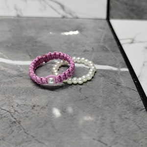 May include: Two rings on a gray surface. One ring is a woven, light purple color with a single white pearl in the center. The other ring is white and made of small, round pearls. The rings are next to each other.