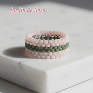 以下が含まれることがあります： ピンクとオリーブグリーンの模様のビーズリング。リングにはピンク色のスクリプトで「Jardin de Rose」の文字と、Instagramのハンドルネーム「@handcraftart.mariam」が記載されています。リングは白い表面に置かれています。