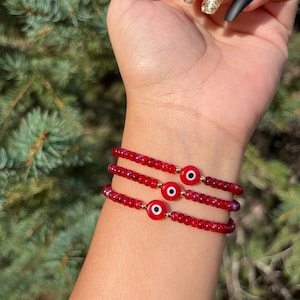 May include: Three red beaded bracelets with gold accents and a red evil eye charm on each bracelet.