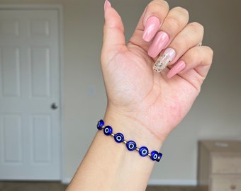 May include: A blue evil eye bracelet with gold beads. The bracelet is on a person's wrist. The person is wearing pink and gold nail polish.