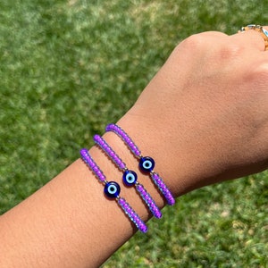 May include: Three purple beaded bracelets with blue evil eye charms. The bracelets are stacked on a person's wrist.