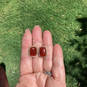 May include: A pair of drop earrings featuring rectangular, smooth, deep red stones set in gold-toned metal frames with a delicate, intricate wirework border. Each earring is suspended from a simple, curved gold-toned hook for secure attachment.