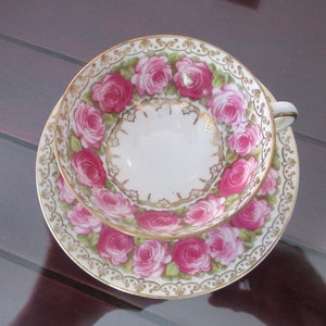 May include: A white teacup and saucer with a gold rim and pink rose floral pattern. The cup and saucer are decorated with gold accents and a delicate floral design.