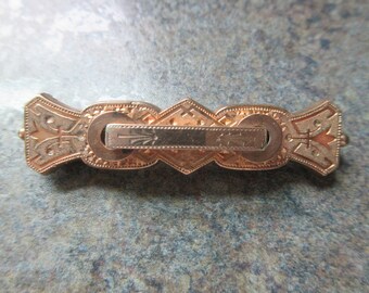 May include: A gold-toned metal brooch with an intricate design. The brooch has a central bar with a decorative pattern and two side pieces with floral motifs.