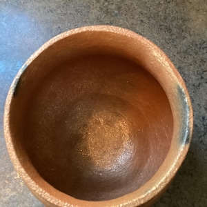 May include: A round, brown ceramic bowl with a textured surface. The bowl has a wide opening and a smooth interior. The exterior has a slightly rough texture, adding to its rustic charm. Ideal for serving or display. The bowl is approximately 15 cm in diameter.