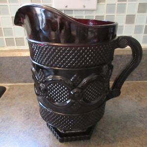 Puede incluir: Una jarra de vidrio de color rojo oscuro con un diseño texturizado y un asa. La jarra tiene una base hexagonal.