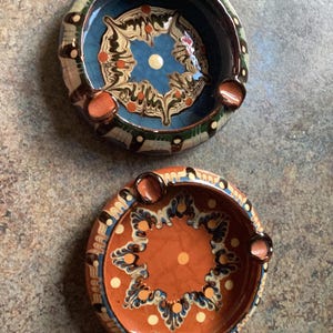 Puede incluir: Dos ceniceros redondos de cerámica con un diseño decorativo. Uno tiene un interior azul con un patrón floral blanco y naranja. El otro tiene un interior naranja con un diseño similar. Cada cenicero tiene tres reposacigarrillos.