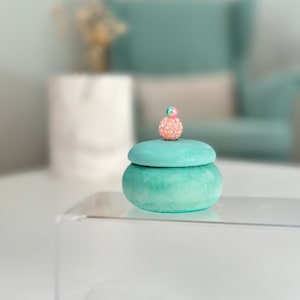 May include: A teal green ceramic trinket box with a round lid. The lid is decorated with a pink and green jeweled ball.