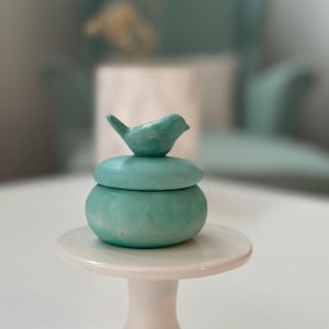 Op de afbeelding: Een kleine, turquoise sieradendoos met een vogelvormig deksel. De ronde doos staat op een witte sokkel. De vogel zit bovenop het deksel en voegt een decoratief tintje toe.