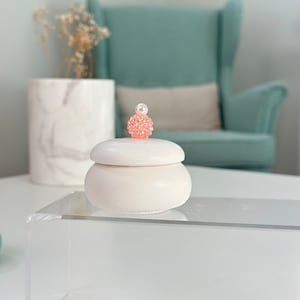 May include: White ceramic trinket box with a pink and white pearl-like lid. The box is sitting on a clear acrylic shelf.
