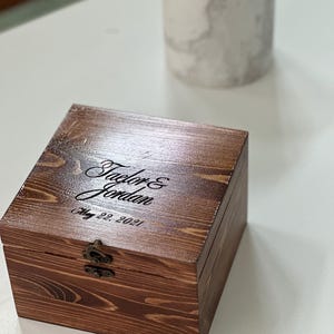 May include: A square, dark-stained wooden box with a hinged lid and a bronze-colored metal latch. The top of the box is engraved with the names Taylor & Jordan and the date May 22, 2021, in a black script font, showcasing a visible wood grain.