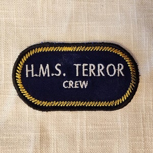Könnte beinhalten: Ein ovaler, bestickter Aufnäher mit dunkelblauem Hintergrund und einem dicken, gelb gestickten Rand. Der Text H.M.S. TERROR ist in weißen Großbuchstaben in der oberen Zeile eingestickt, darunter steht das Wort CREW in weißen Buchstaben.