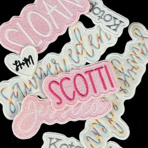 May include: A collection of embroidered name patches in various colors, including pink, white, and gray. The patches feature names like "Sloan," "Scotti," and "Jessica" in a cursive font. A heart-shaped patch with "H+M" is also present.