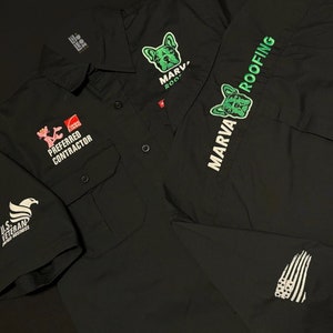 May include: Black work shirt with embroidered logos. The logos include "Marva Roofing", a pink panther with "Preferred Contractor", a French bulldog with "Marva Roofing", and a U.S. Veteran Owned Business eagle.