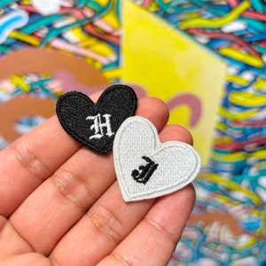 May include: Two felt heart-shaped patches with embroidered letters. The black patch has a white "H" and the white patch has a black "J".