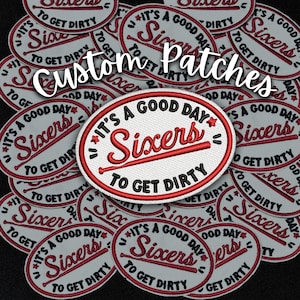May include: A close-up of several white oval-shaped patches with red and black embroidery. The patches feature the text "It's a good day" and "Sixers" with a baseball bat graphic. The text "To get dirty" is also included on the patches.