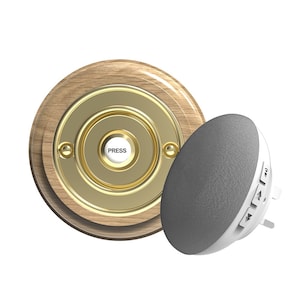 Puede incluir: Un botón de timbre redondo de madera con una placa frontal dorada y la palabra "PRESS" en el botón. También se muestra un pequeño altavoz redondo gris con botones de control de volumen.