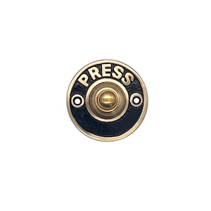 Puede incluir: Un timbre de puerta redondo de latón con una cara negra y la palabra "PRESS" en letras doradas. El timbre tiene un botón dorado en el centro.