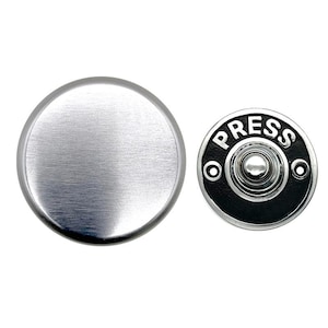 Può includere: Un pulsante per campanello rotondo in argento spazzolato con un pulsante rotondo nero che dice "PRESS" in lettere argentate.