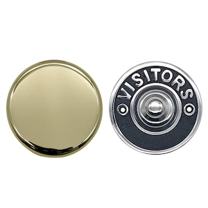 May include: Two doorbell buttons. One is a shiny gold-colored, circular button. The other is a black button with the word "VISITORS" in white lettering around the edge. A silver button is in the center.
