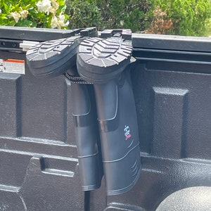 May include: Two black rubber boots hanging from a truck bed rail using a boot holder. The boots have a black strap with white text that says "Hunter" on the right boot.