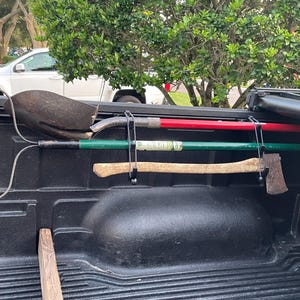 Peut inclure: Un plateau de camion noir avec une pelle, une hache et un râteau accrochés à des crochets. Les outils sont fixés avec des cordons élastiques.