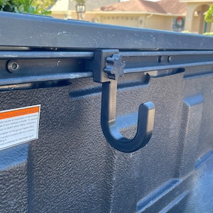 May include: A black plastic hook with a star-shaped knob attached to a black metal track. The hook is designed to be used in a truck bed.